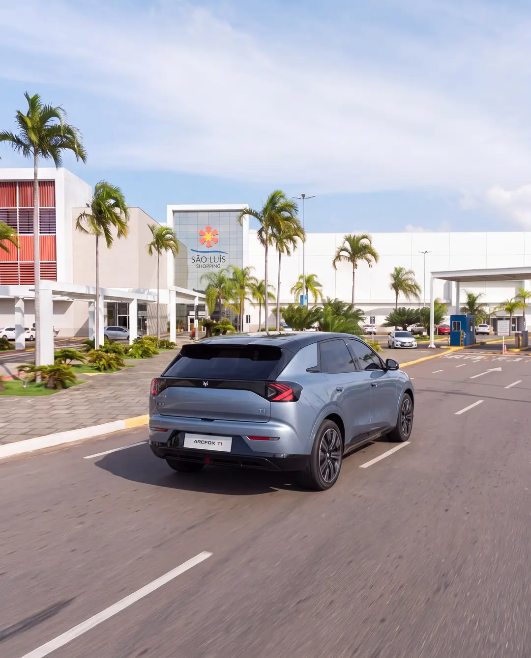 Arcfox T1 entrando no São Luís Shopping, visto de trás, entre palmeiras, com o letreiro do shopping na fachada